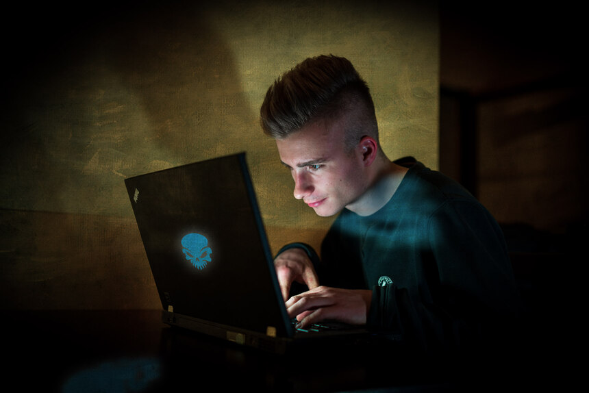 Eine junge männliche Person sitzt im Dunkeln vor einem leuchtenden Laptop.