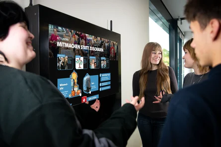 mehrere junge Personen stehen vor einem Display und reden miteinander. Auf dem Bildschirm steht als Überschrift "Beteiligung. Mitmachen statt zuschauen"