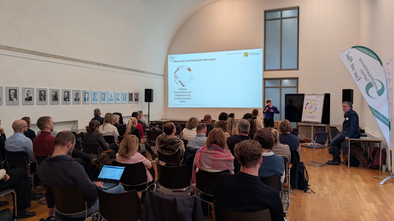 Eine Frau vor einem Publikum einen Vortrag. Ihre Präsentation wird an die Wand projiziert.