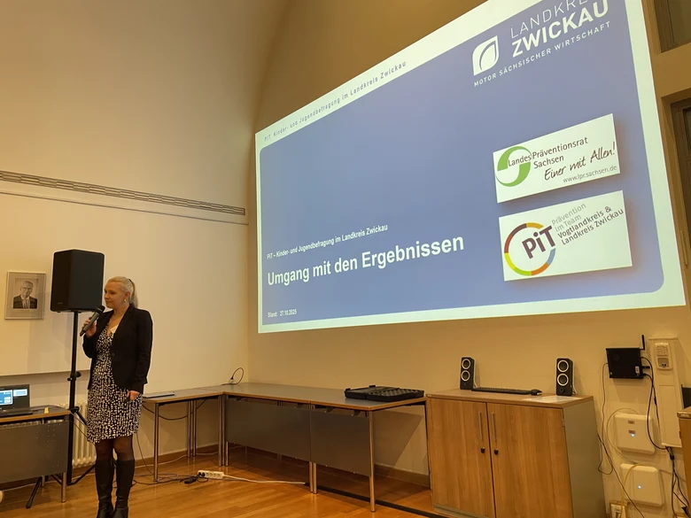 Eine Frau steht mit einem Mikrofon in der Hand. Dahinter wird ihre Präsentation an die Wand projiziert.