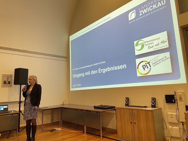 Eine Frau steht mit einem Mikrofon in der Hand. Dahinter wird ihre Präsentation an die Wand projiziert.