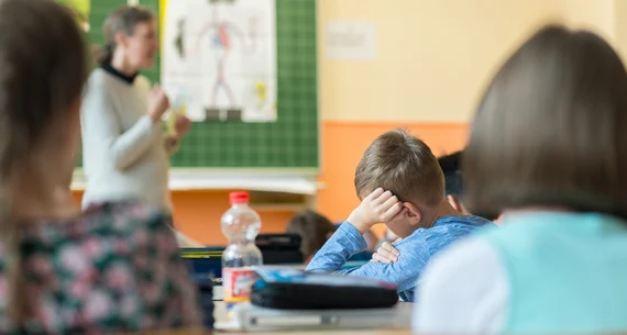 Kinder einer Schulklasse sitzen und hören zu, wie unscharf im Hintergrund eine erwachsene Person etwas erzählt.
