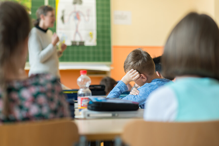 zeigt Klassenzimmer, drei Kinder von Hinten, ein Junge im Fokus aufgestützt auf den Tisch, eine Lehrperson im Hintergrund