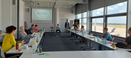Elf Männer und Frauen sitzen in einem großen Raum und schauen auf eine Präsentation. Rechts kann durch das Fenster ein stehendes Flugzeug beobachtet werden.