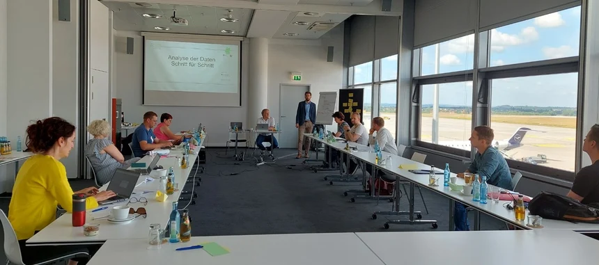 Elf Männer und Frauen sitzen in einem großen Raum und schauen auf eine Präsentation. Rechts kann durch das Fenster ein stehendes Flugzeug beobachtet werden.