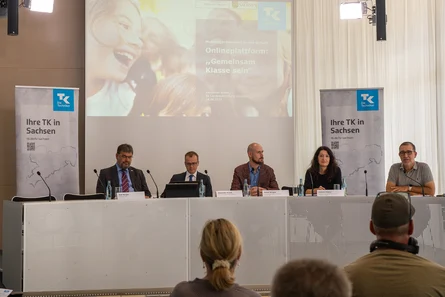 Fünf Personen, vier Männer und eine Frau, sitzen an einem Konferenztisch mit Blick zum Podium. Hinter ihnen ist eine Präsentation der Techniker Krankenkasse zum Thema "Onlineplattform: 'Gemeinsam Klasse sein'".