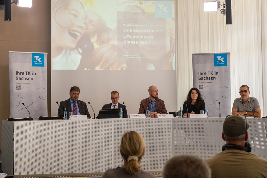 Fünf Personen, vier Männer und eine Frau, sitzen an einem Konferenztisch mit Blick zum Podium. Hinter ihnen ist eine Präsentation der Techniker Krankenkasse zum Thema "Onlineplattform: 'Gemeinsam Klasse sein'".