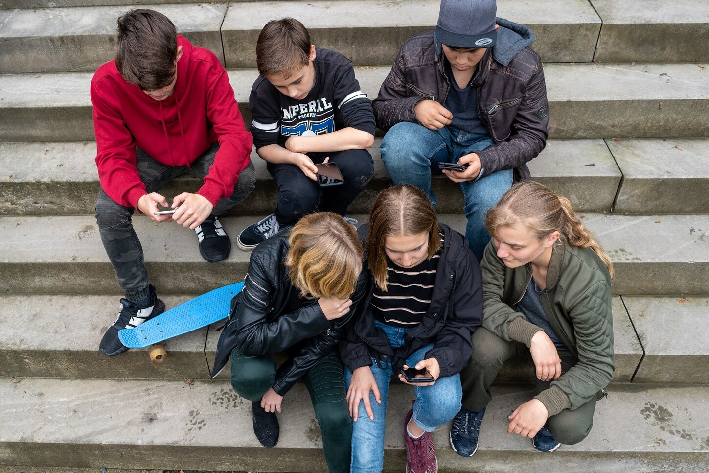 Mehrere Kinder und Jugendliche sitzen auf Treppestufen. Jeder hält ein Handy in der Hand und jeder schaut auf sein Handy oder auch auf das Handy des anderen.