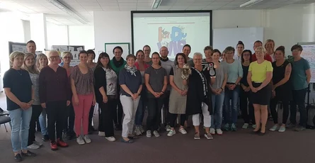 Eine große Gruppe steht in einem Raum und schaut in die Kamera. Eine Frau hält zwei Figuren in den Händen. Hinter den Personen ist wird eine Präsentation projiziert.