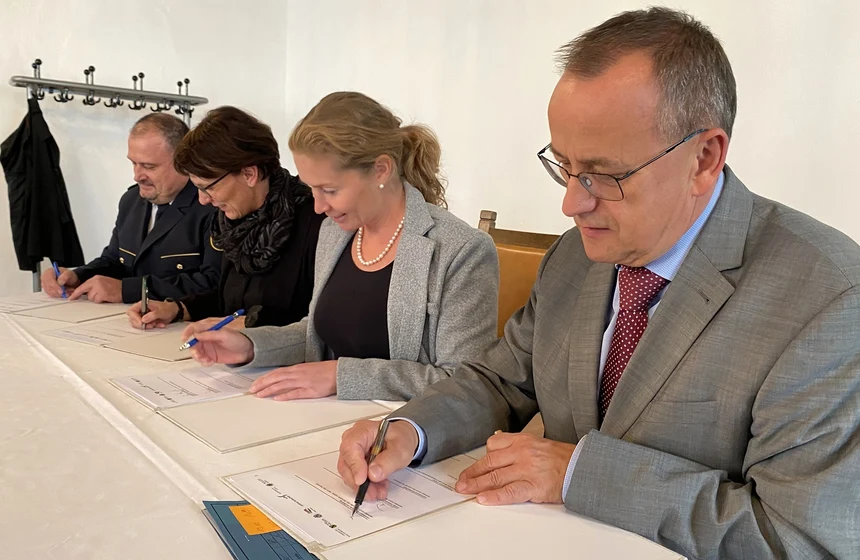 Vier Personen, zwei Männer und zwei Frauen, sitzen an einem Tisch und unterzeichnen Dokumente.