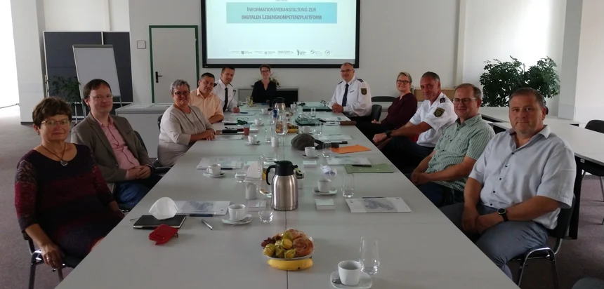 Elf Männer und Frauen sitzen an einem Tisch und schauen in die Kamera. Hinter ihnen wird eine Präsentation abgespielt.