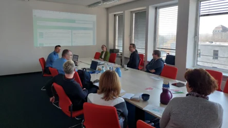 Acht Männer und Frauen sitzen an einem Tisch. An die Wand wird eine Präsentation projiziert.