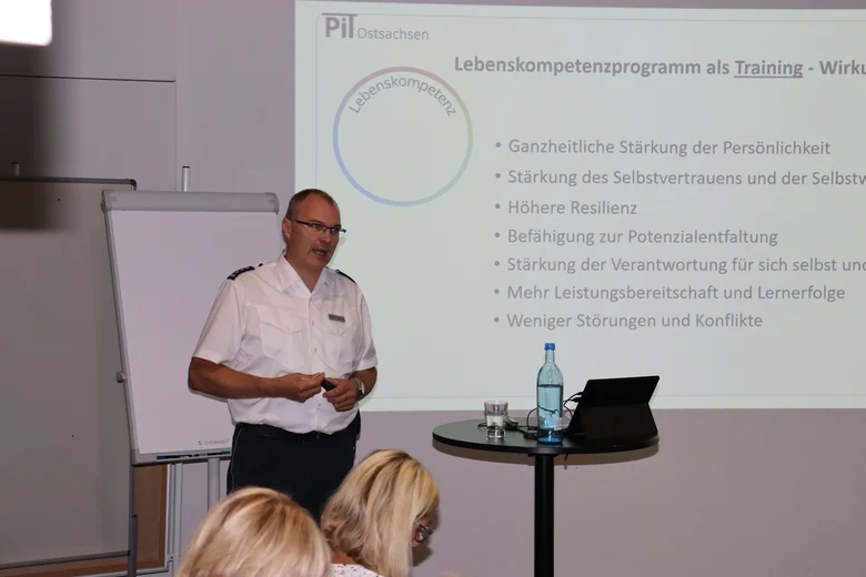 Ein Mann steht vor einer Wand, auf die eine Präsentation projiziert wird. Dabei hält er einen Vortrag.