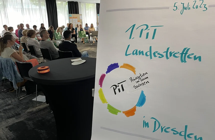 Im Hintergrund sitzen viele Menschen. Im Vordergrund steht rechts ein Aufsteller, auf dem "1. PiT Landestreffen" steht.
