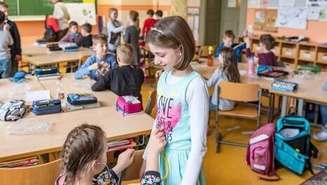 Im Hintergrund ist eine Schulklasse mit jüngeren Kindern. Im Vordergrund sind zwei Mädchen. Das Mädchen auf der linken Seite hört mit einem Stethoskop den Bauch des anderen Mädchens ab.