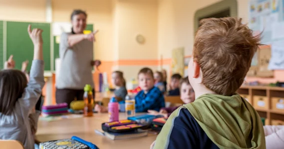 Im Klassenraum sitzen junge Kinder. Im Vordergrund sieht man ein Kind von hinten und im Hintergrund eine Lehrkraft sowie weitere Kinder.