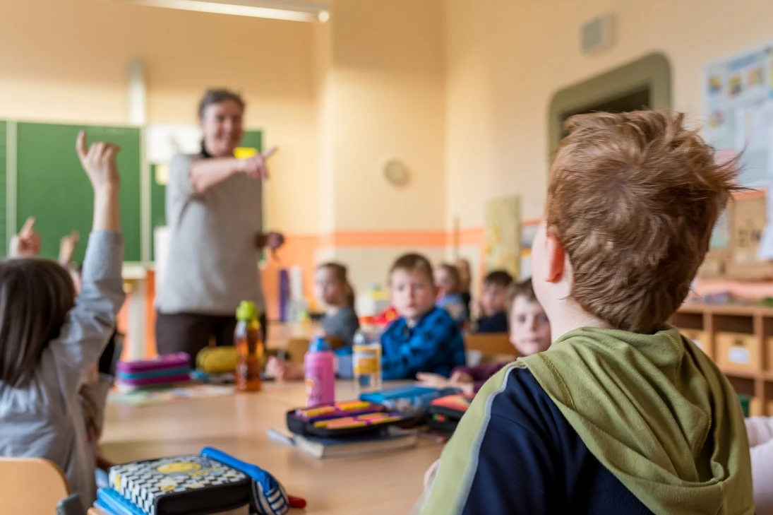 Im Klassenraum sitzen junge Kinder. Im Vordergrund sieht man ein Kind von hinten und im Hintergrund eine Lehrkraft sowie weitere Kinder.