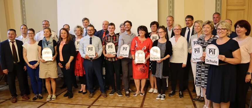 26 Personen stehen zusammen in mehreren Reihen und schauen nach vorne. Einige von ihnen halten Tafeln mit der Aufschrift "PiT-Ostsachsen-Schule".