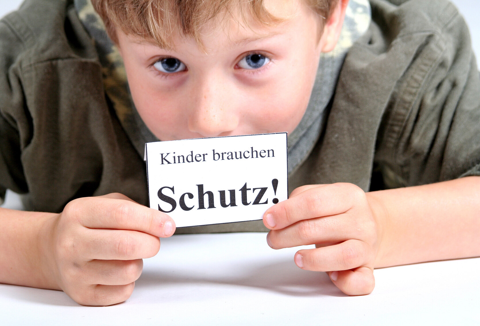 Ein Junge mit olivgrünem Oberteil liegt auf einer weißen Fläche. Dabei sieht man nur seinen Kopf, Schultern und Hände. In den Händen hält er einen weißen Zettel mit den Worten "Kinder brauchen Schutz!" und verdeckt damit teilweise sein Gesicht.