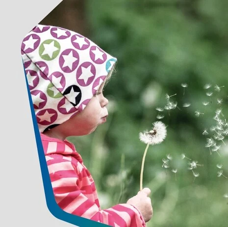 Auf der linken Seite ist ein kleines Kind im Profil, welches eine Pusteblume in der Hand hält und pustet. Das Kind trägt eine weiße Mütze mit bunt umrandeten Sternen und eine rosafarbene gestreifte Jacke.