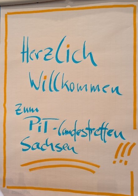 Auf einem Flipchart steht ein handgeschriebener Begrüßungstext in türkisblauer Schrift, um-rahmt von einem einfachen orangefarbenen Rand. Zu lesen ist: „Herzlich Willkommen zum PiT-Landestreffen Sachsen“. Unten rechts stehen drei orange Ausrufezeichen und