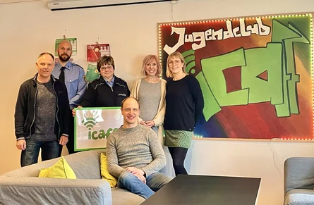 Fünf Personen, zwei Männer und drei Frauen, stehen in einem Raum vor einem Plakat mit der Aufschrift "Jugendclub icaff" und lächeln. Eine sechste Person, ein Mann, sitzt vor ihnen auf einem Sofa.