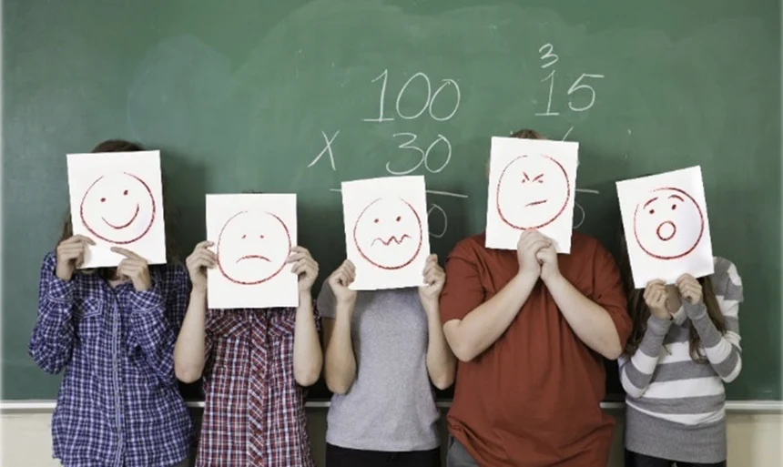Fünf junge Personen stehen vor einer grünen Tafel und halten sich weiße Zettel vor die Gesichter. Darauf sind symbolhafte Gesichter gemalt, die verschiedene Gefühle ausdrucken.