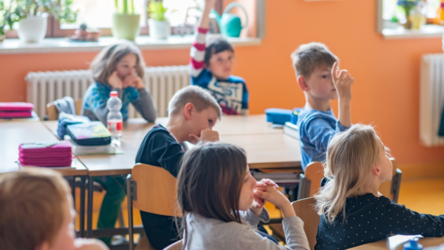 Junge Schülerinnen und Schüler sitzen in einem Klassenzimmer an ihren Tischen und schauen nach vorne. Ein Junge hebt die Hand.