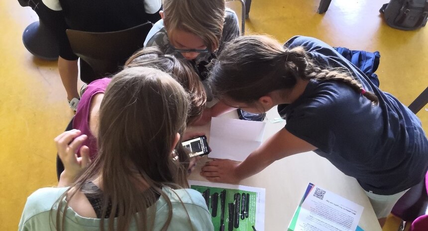 Kinder von oben fotografiert beugen sich in einen Klassenzimmer über eine Schulbank und schauen auf ein Smartphone