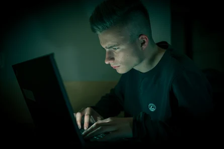 Eine junge männliche Person sitzt im Dunkeln vor einem leuchtenden Laptop.