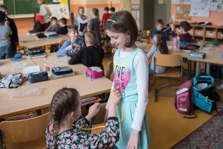 Im Hintergrund ist eine Schulklasse mit jüngeren Kindern. Im Vordergrund sind zwei Mädchen. Das Mädchen auf der linken Seite hört mit einem Stethoskop den Bauch des anderen Mädchens ab.