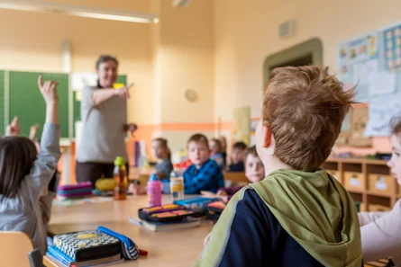 Im Klassenraum sitzen junge Kinder. Im Vordergrund sieht man ein Kind von hinten und im Hintergrund eine Lehrkraft sowie weitere Kinder.